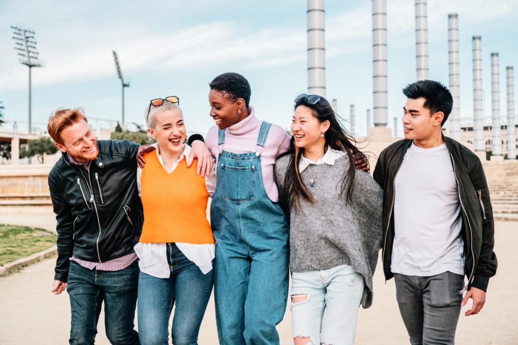 Five young adult friends meeting hugging and laughing outdoors.