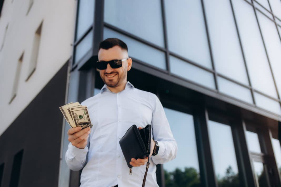 Young business man standing leaning on wall wearing eyeglasses on the city street counting money